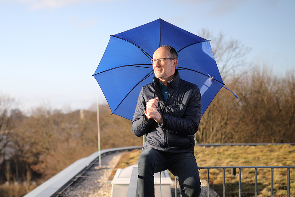 Mann mit blauem Regenschirm sitz bei Sonnenschein auf einer Absperrung.