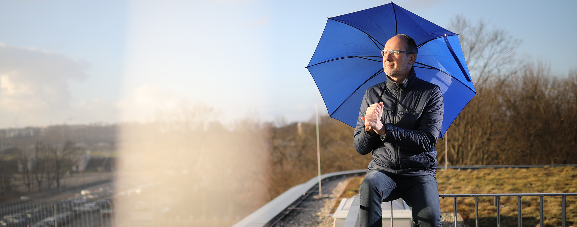 Mann mit blauem Regenschirm sitzt bei Sonnenschein auf einer Absperrung.