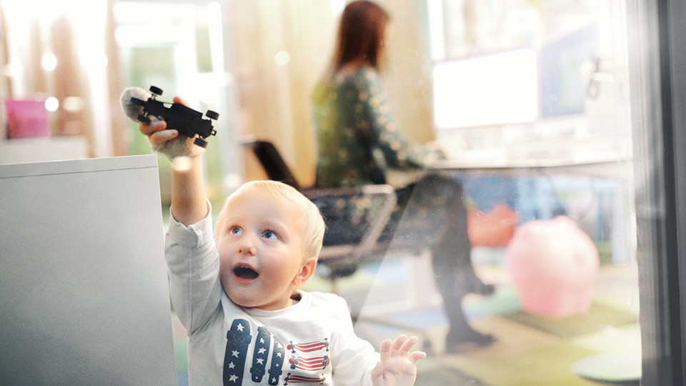 Kinder spielt mit Spielzeugauto an einer Fensterscheibe, im Hintergrund arbeitet eine Frau im Eltern-Kind-Büro.